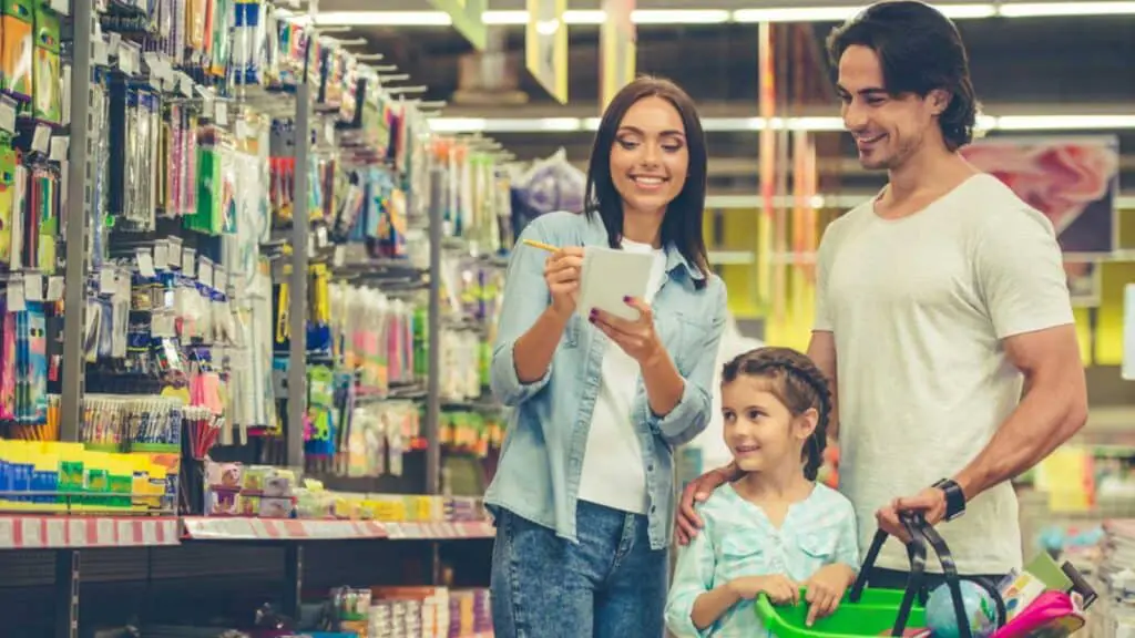 Beautiful Young Parents And Their Cute Little Daughter Are Smiling While Choosing School Stationery In The Supermarket. Mom Is Making Notes In The List