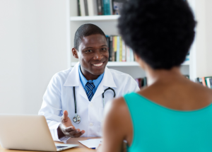 Can You Trust Osteopathic Doctors? &Raquo; Photo Of A Smiling Black Male Doctor Sitting At His Desk Talking To A Female Patient Who Is Facing Him Canva 1 300X215 1