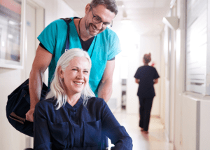 Tips And Strategies For Coming Home From The Hospital &Raquo; Photo Of A Smiling Woman Leaving The Hospital Being Pushed An Aide Who Is Carrying Her Suitcase Canva 1 300X214 1