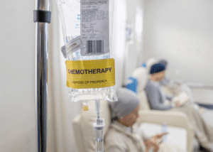 6 Important Things To Know About Chemotherapy &Raquo; Photo Of A Bag Of Chemotherapy Medication Hanging From A Pole With Blurred Patients Sitting In The Background Canva 1 300X214 1