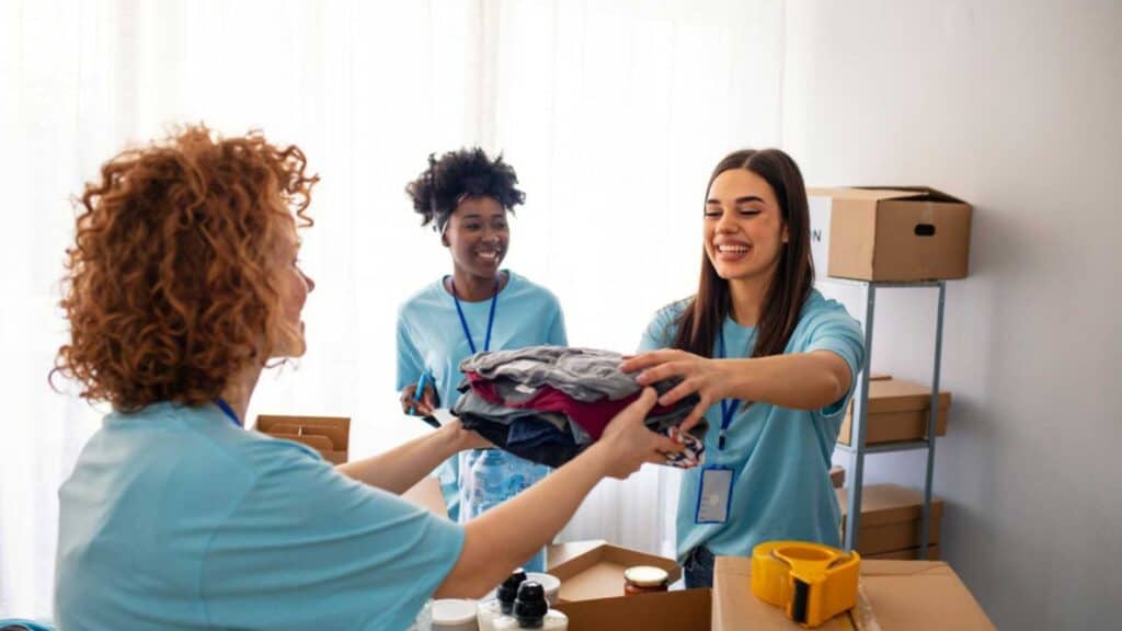 Volunteers Collecting Food Donations In Warehouse. Team Of Volunteers Holding Donations Boxes In A Large Warehouse. Volunteers Putting Clothes In Donation Boxes, Social Worker Making Notes Charity