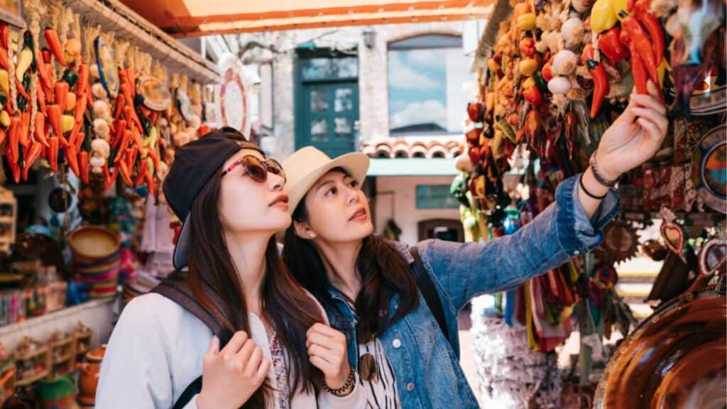 Pretty Backpackers Buying Chili Decorations In The Traditional Market. Two Travelers Shopping In A Colorful Vendor. Mexican Decorations Shop In Usa.