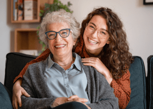 Caregiving Tips For Managing Incontinence In Seniors &Raquo; Photo Of A Young Woman With A Big Smile Hugging Her Grandmother From Behind Who Also Has A Big Smile While They Sit On A Sofa Canva 1 300X214 1