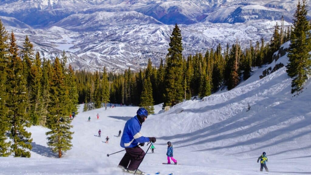 Senior Male Skier Skiing In Colorado Ski Resort Near Aspen, Colorado, On Nice Winter Day; More Skiers And Snowboarders Skiing Around; Woods And Mountains In Background