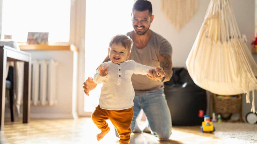 Happy Father Helping Little Son Walking In Living Room