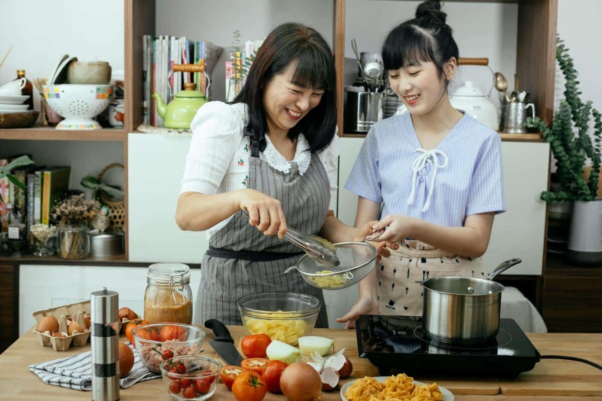 Laughing Asian Mother And Daughter Preparing Pasta In Kitchen