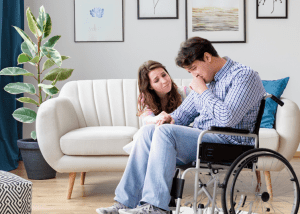 Managing Anxiety And Depression After A Traumatic Injury &Raquo; Photo Of A Young Woman Kneeling At Her Husbands Side As He Sits In A Wheelchair Looking Sad And Anxious Canva 1 300X214 1