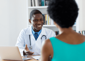 Can You Trust Osteopathic Doctors? &Raquo; Photo Of A Smiling Black Male Doctor Sitting At His Desk Talking To A Female Patient Who Is Facing Him Canva 1 300X215 1