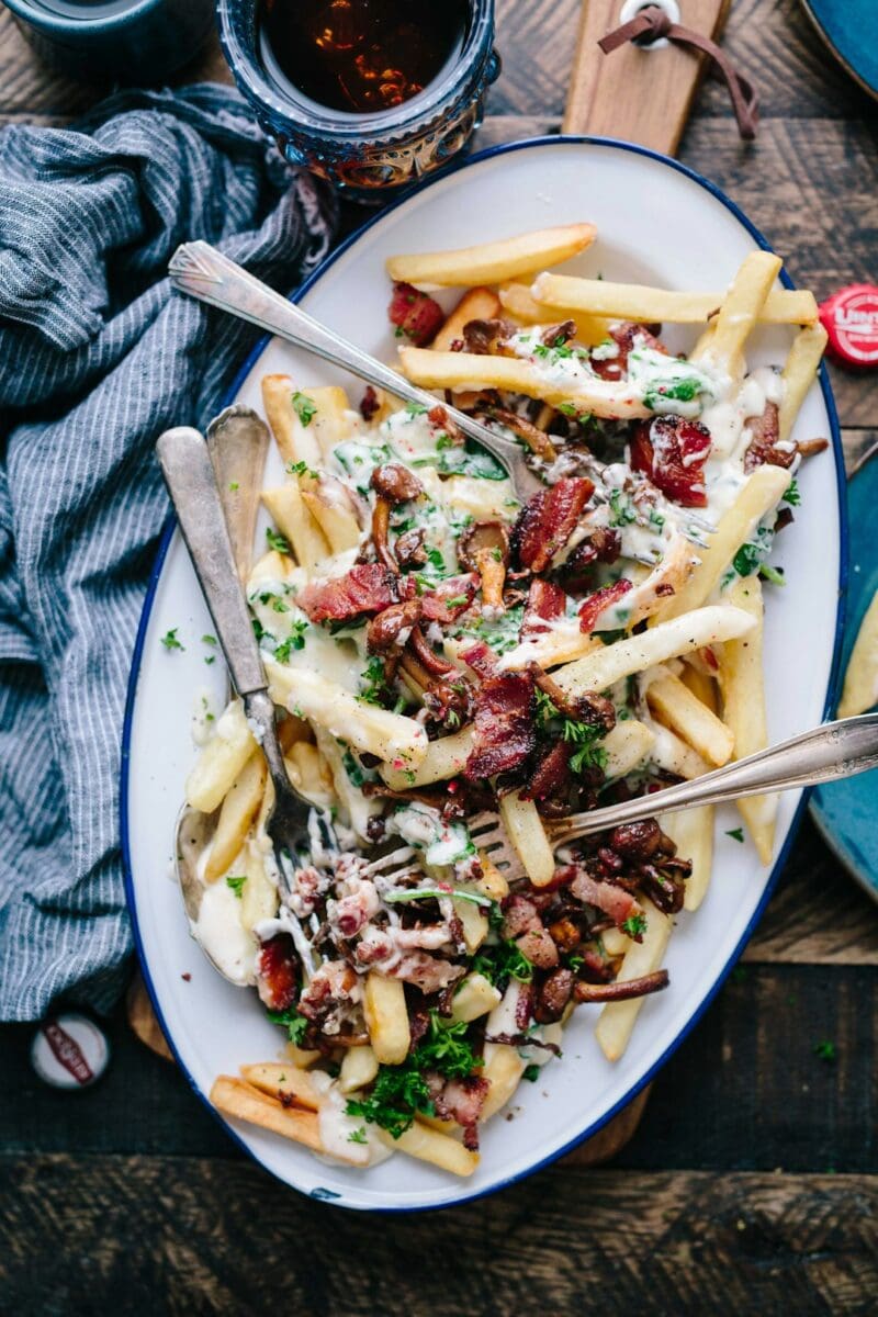 Bacon Strips And Melted Cheese Topped Fries On Oval White And Blue Platter With Gray Stainless Steel Forks